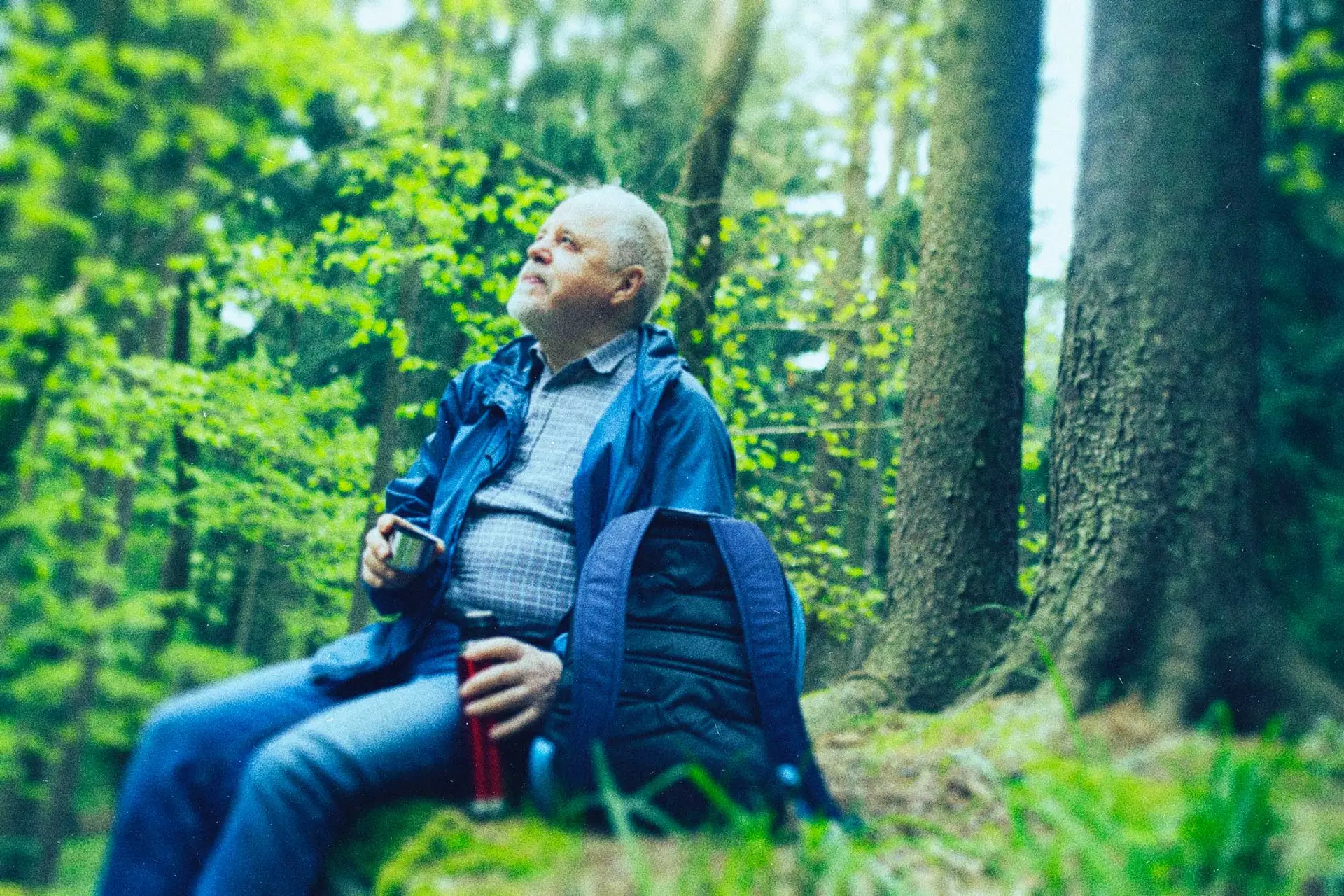 mature man in the woods with a coffee thermos and backpack, sitting on a grassy ledge looking up to the sky