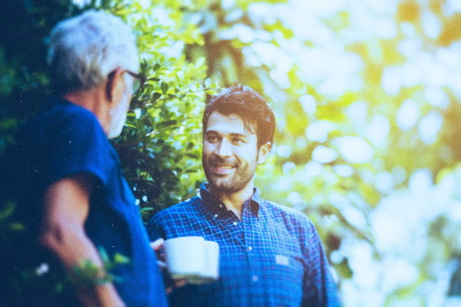 senior man and younger man talking with coffee cups near bushes of trees and the sun beaming