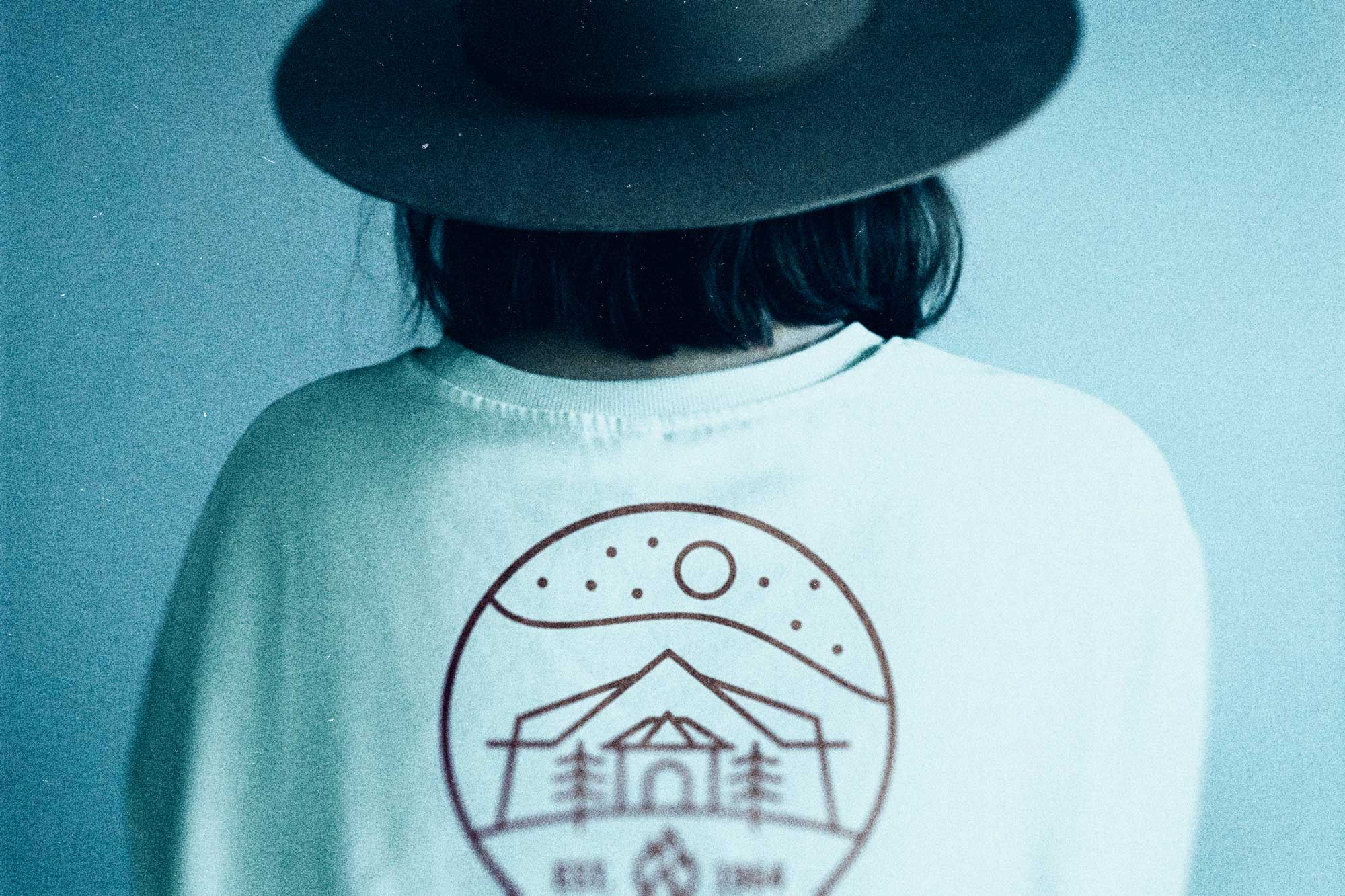 Backside of a woung woman wearing a hat and a Club T-shirt with the logo printed in rust-orange on the back